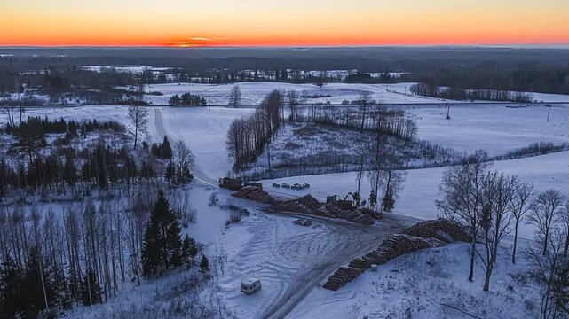 A static aerial hyperlapse of heavy-duty loaders and forestry cranes stacking timber in the snow at golden hour sunset, capturing the winter wood-processing workflow.