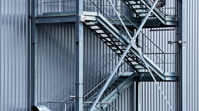 Drone rises along exterior of industrial warehouse revealing zigzagging galvanized steel staircases, landings and railings against corrugated metal facade in cool blue-grey tones
