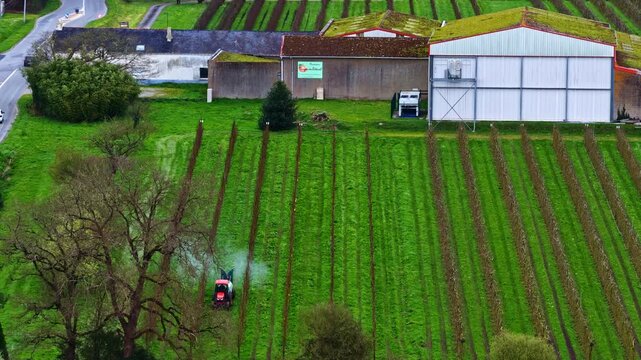 Drone captures red tractor spraying pesticide along lush green rows of young fruit trees in French orchard near farm warehouse and road roundabout in spring