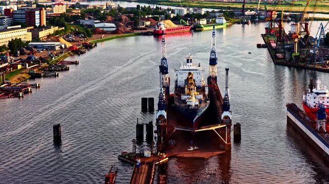 Large cargo ship in port channel lined with cranes docks and industrial buildings, aerial