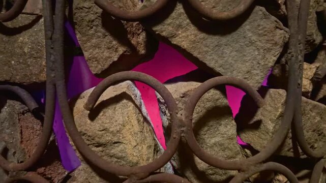 Close-up shot of ornate rusty wrought iron gate with circular scrolls against rough stone wall, vivid pink light glowing through gaps