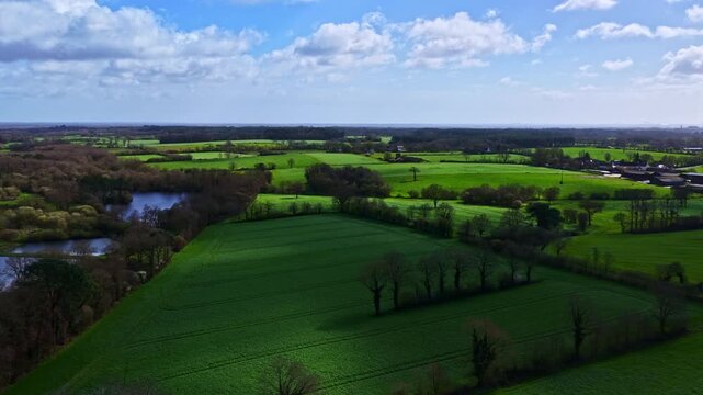 Aerial time lapse captures moving cloud shadows sweeping over vibrant green patchwork fields, rolling hills, hedgerows, and ponds in scenic English countryside