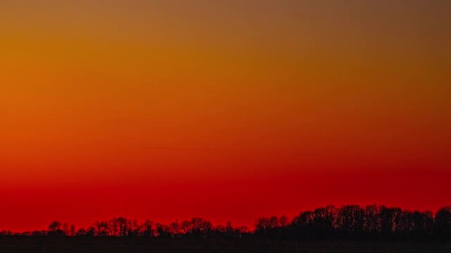 Hazy sky glows orange and red above a dark tree line at dusk in timelapse