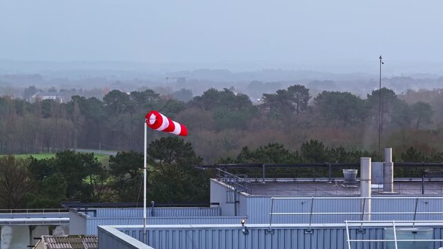Static shot of an industrial building rooftop in Latvia with a red and white windsock indicating strong wind against a hazy forest and distant town background
