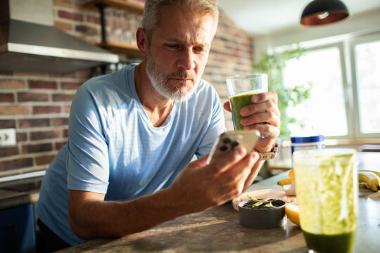 Middle-aged man drinking green smoothie at home kitchen while checking smartphone