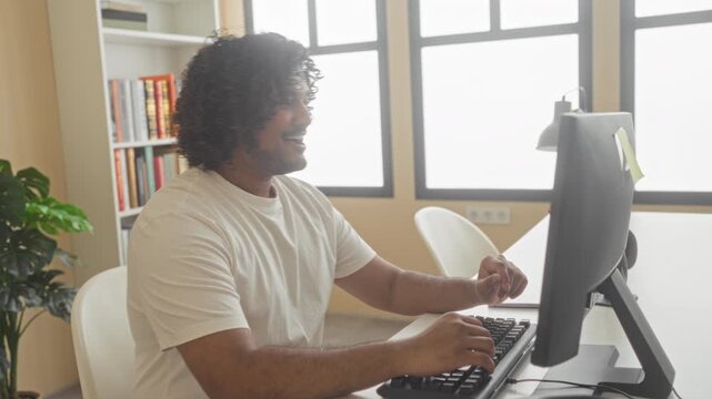 Man with bare arms leans back hands behind head at computer desk in building with bookshelf, monitor and plant, reclining in chair with eyes closed; short break relaxed.
