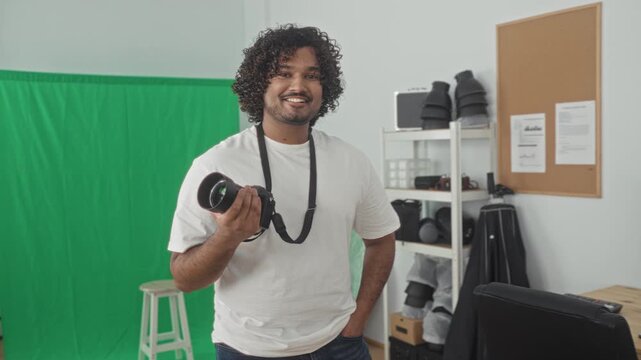Man holding camera and smiling, hand on hip in studio with green screen and lighting gear; creative confidence.