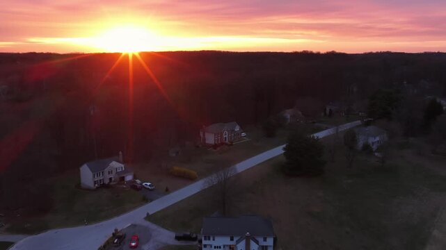 Suburban cul de sac at sunset with warm sunburst over quiet residential homes in America, showing peaceful rural neighborhood with houses and open landscape during calm evening. Aerial flyover shot.