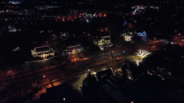 Aerial night view of residential neighborhood decorated with Christmas lights, glowing houses and quiet streets. Festive winter atmosphere in United States during Christmas season. Approaching shot.