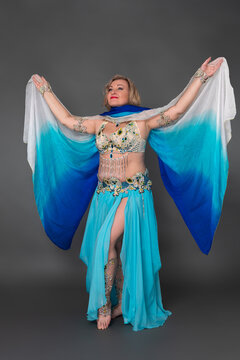 Mid aged adult woman fashion model dressing in dancing stage costume - bra top, blue maxi skirt with slit, pareo and she is not wearing any footwear, standing in full length against gray background