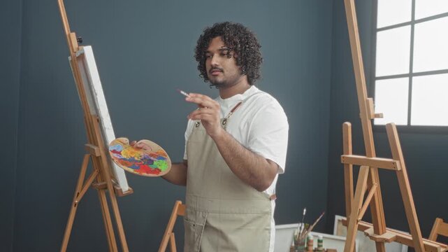 Man painter holds paintbrush and palette while working on canvas in studio; concentration creativity.