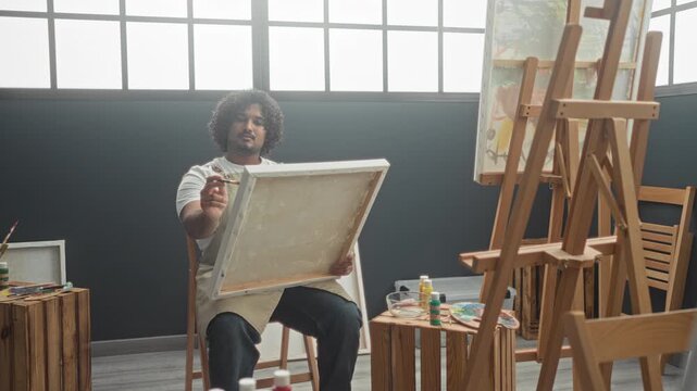 Young indian man painter in apron sits on stool and holds paintbrush to canvas with palette and easel in studio; concentration creativity.