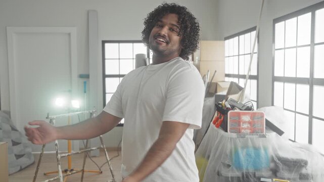 Man with arms crossed in a white t shirt among packed moving boxes, ladder, bubblewrap and a toolbox in a building; moving day pride.