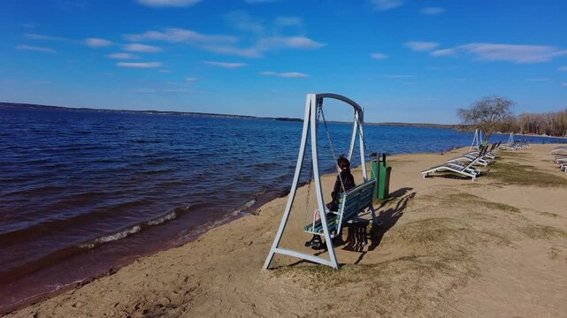 A person swings gently by the water at a beach. The sun shines bright, and clouds drift in the blue sky. Nearby are empty chairs and trees along the shore.