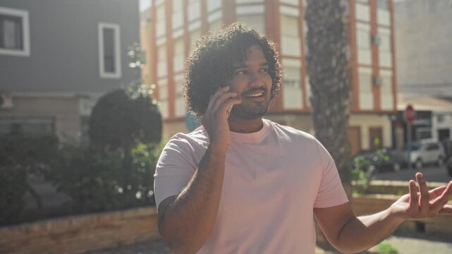 Man holding smartphone to ear and gesturing with hand on a sunny street near a building; casual chat contentment.