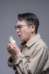 Asian man with glasses blowing his nose with a tissue, suffering from a cold, allergy, or PM 2.5 pollution symptoms against a grey studio background, portrait studio shot © whyframeshot
