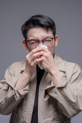 Asian man with glasses blowing his nose with a tissue, suffering from a cold, allergy, or PM 2.5 pollution symptoms against a grey studio background, portrait studio shot © whyframeshot
