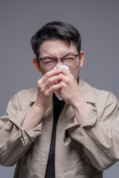 Asian man with glasses blowing his nose with a tissue, suffering from a cold, allergy, or PM 2.5 pollution symptoms against a grey studio background, portrait studio shot