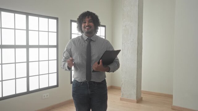 Man holding black folder gestures with open hand and points with other hand in empty building interior, smiling and presenting the space; confidence trust.