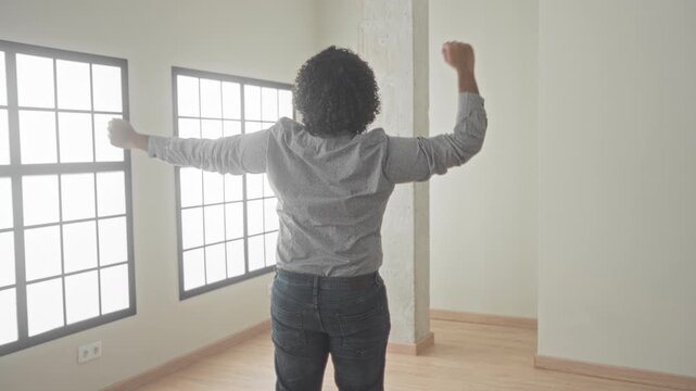 Man with curly hair raising arms while inspecting an empty house interior with large windows, wooden floor and concrete column in a building; homeownership confidence.