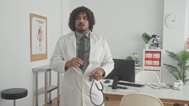 Young indian doctor holding stethoscope, hand to mouth gesture beside desk, computer, anatomy poster and microscope in clinic building; concern care duty.