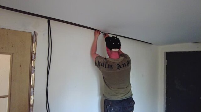 A man works on the ceiling, installing a suspended ceiling. He uses tools and stands on a ladder to reach the required area. The photo shows a simple room with a single wall.