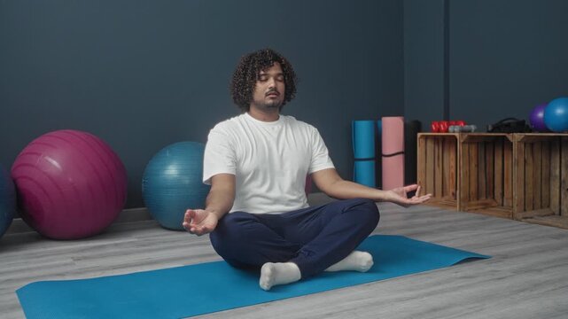Man seated crosslegged on blue mat with hands in gyan mudra meditating in studio; mindfulness serenity.