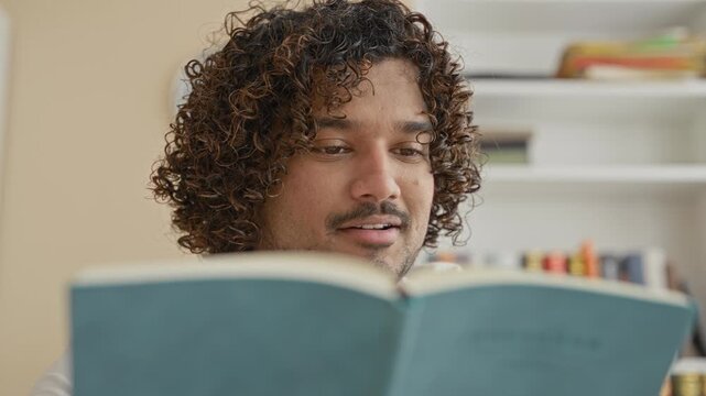 Young indian man reading teal book, hand on chin while turning page in a building with bookshelf and wall clock visible; contemplation.