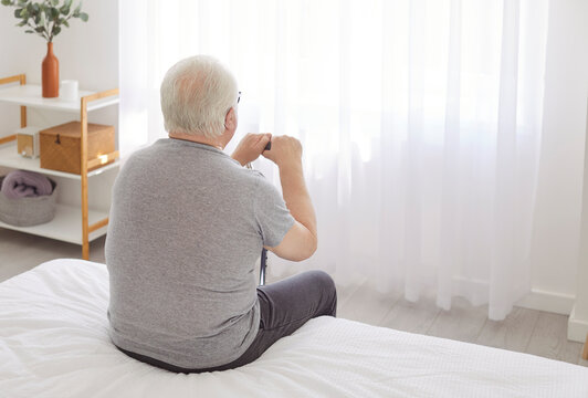 Senior old man alone at home on bed, rear view, lonely pensive grandfather, elderly person retired patient in retirement house, sitting thinking in comfortable nursing home bedroom, old age home