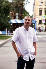Fototapeta premium A fair-skinned man in a white shirt and dark jeans stands in the middle of a St. Petersburg street, glancing upward. A blue trolley bus and other buildings are visible in the background.
