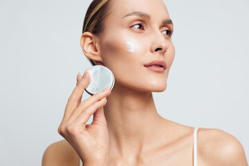 Woman skincare face cream cotton pad beauty hygiene portrait with clean studio light, she applies moisturizer to her cheek, calm expression, closeup beauty routine for skin care © SHOTPRIME STUDIO