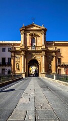 Obraz premium An ornate, arched stone gateway under a clear blue sky, leading into a city street with buildings on either side. The structure has a cross