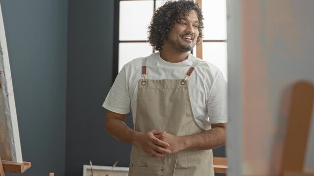 Smiling young indian painter man in apron with arms crossed beside easel and canvas by window in studio; calm focus.