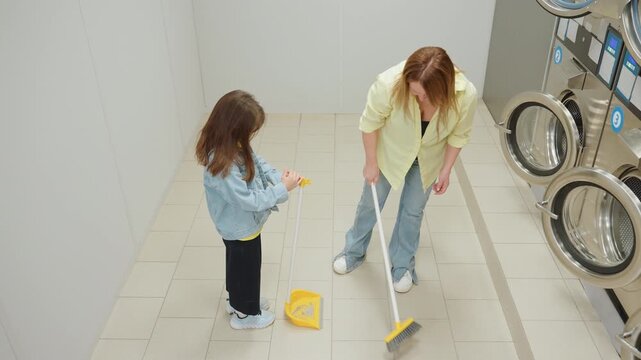 Overhead view shows shop owner sweeping tiled laundromat floor while toddler helper holds dustpan near stainless washers, teamwork routine, family hygiene focus, interior, denim jeans, yellow tools