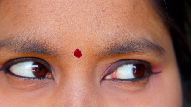 Extreme close-up of an Indian woman's eyes with pink eyeshadow, black eyeliner, and a red bindi, looking left and right.
