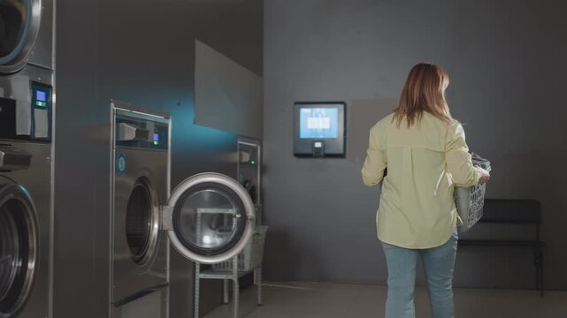 Female worker carries basket of clothes, walks away from industrial washer inside laundromat, machines in row, piles of towels visible, routine service, clean facility, daily chores, smooth operation