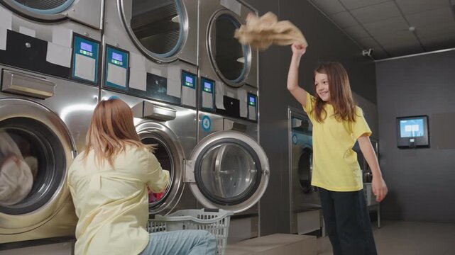 Mother with kid packing clothes into washer at laundromat, woman kneels by open stainless machine, child gestures while basket of towels waits, sorting textiles, clean service routine inside
