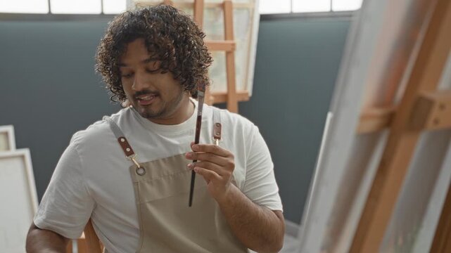 Man painter holding paintbrush and gesturing with hand in studio by easel and canvas while selecting brushstroke technique; concentration creativity.