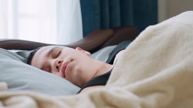 Close up boy resting on pillow with eyes closed wearing black singlet looking restless under beige blanket in bedroom showing discomfort during sleep peaceful yet uneasy