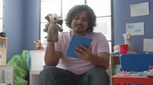 Man holding tablet and plush elephant puppet while gestures to toy in kindergarten building; nurturing warmth.
