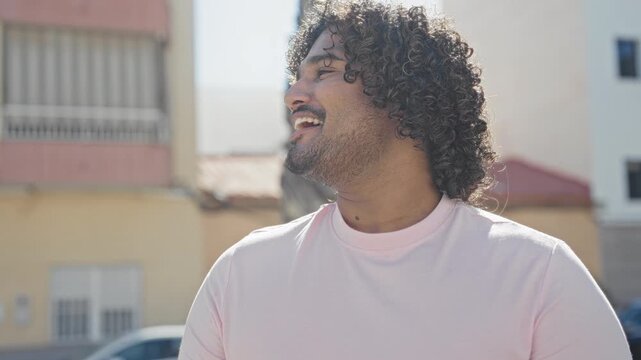 Man smiling and looking up with face visible, curly hair and beard, wearing pink t shirt in bright sunlight on a residential street with buildings and parked car; joy.