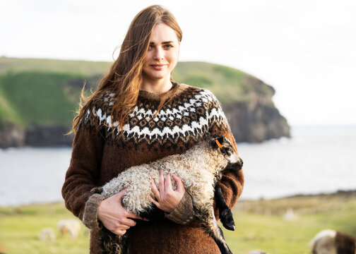 A girl stands with a lamb in her arms in Icelandic sweater.