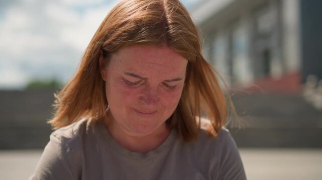 Emotional woman with windblown hair stands under bright clear sky, eyes lowered in thought, gentle sunlight highlighting face, conveying deep reflection