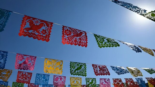 Papel picado mexicano ondeando bajo un cielo azul brillante y sol intenso; decoraci&oacute;n festiva para Cinco de Mayo, herencia cultural y celebraciones tradicionales vibrantes.
