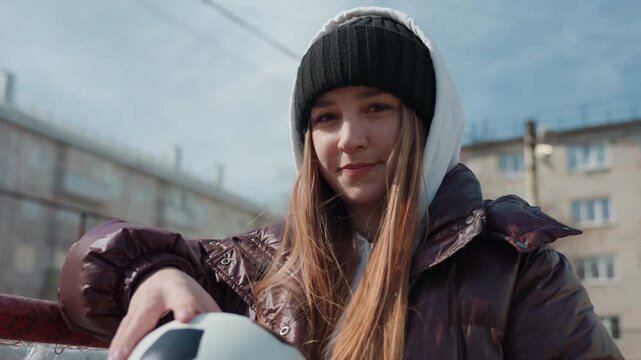 smiling caucasian teen with soccer ball leaning on railing, hoodie and long hair catching light, relaxed urban court backdrop, warm smile and casual posture, residential buildings in distance,