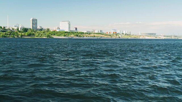 A scenic view of a modern European city skyline from across a wide, deep blue river. The water has small waves, and the distant bank is covered in green trees with high-rise buildings