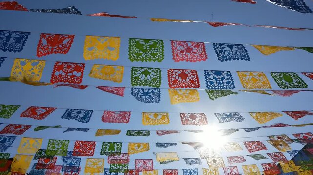 Papel picado mexicano tradicional ondeando bajo el sol y cielo azul. Decoraci&oacute;n festiva para Cinco de Mayo y D&iacute;a de Muertos. Ideal para contenido de viajes, cultura y festividades.