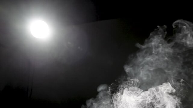 Soft white smoke billows and curls across a dark background, illuminated by a bright glowing light source on the left, creating dramatic contrast and atmospheric depth with copy space