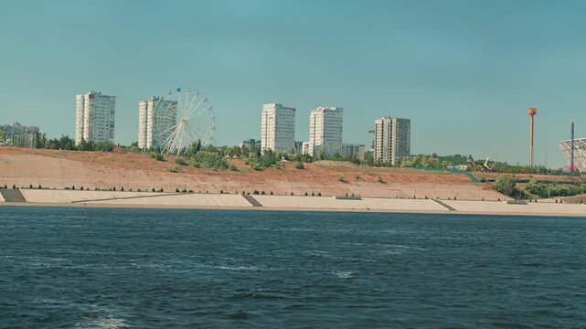 Panoramic view of a modern city skyline with residential buildings and a ferris wheel. The city is on a high, sandy embankment next to a wide, calm river under a clear sky. Calm mood
