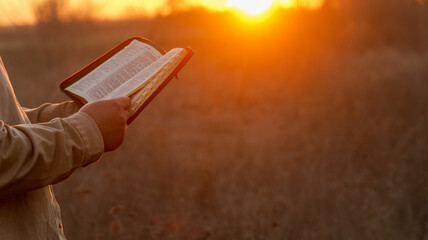 Person reading a Bible outdoors during a warm sunset with golden light illuminating the scene